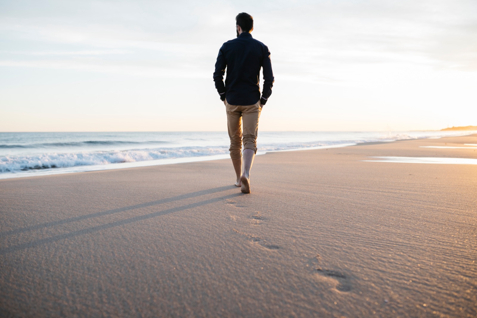 A man walking along the shoreline at sunset, footprints behind him